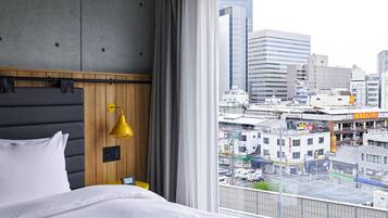 A hotel room with a bed, a wooden headboard, a yellow lamp, and a city view through large windows.