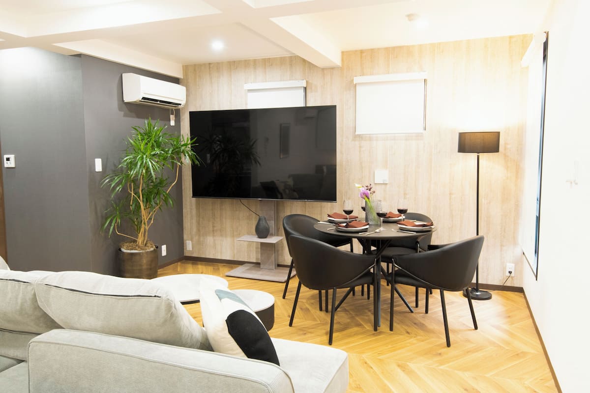 A modern living room with a sofa, a dining table with chairs, a large flat-screen TV, and a potted plant.