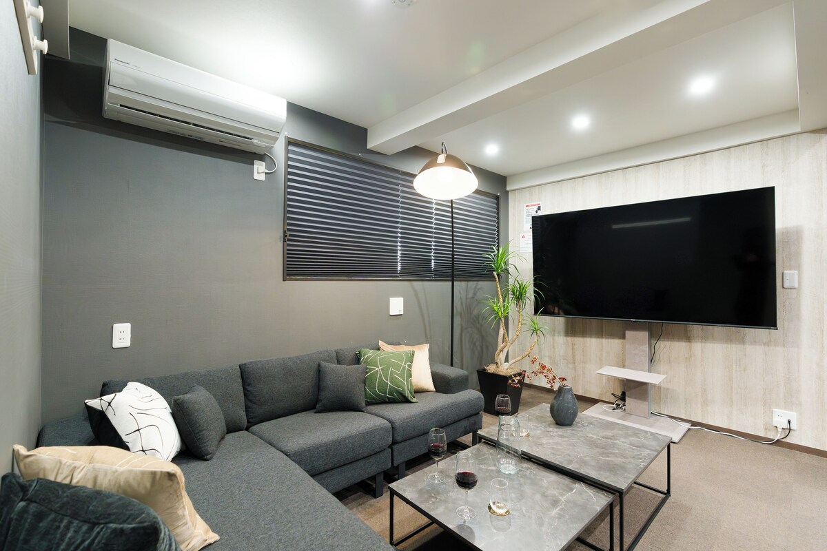 Una sala de estar moderna con un sofá seccional, un televisor de pantalla plana montado en la pared, una mesa de centro y una planta en maceta.