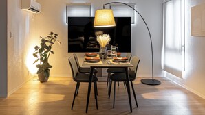 Een eetruimte met een tafel voor vier personen, een vloerlamp, een potplant en een grote flatscreen televisie.