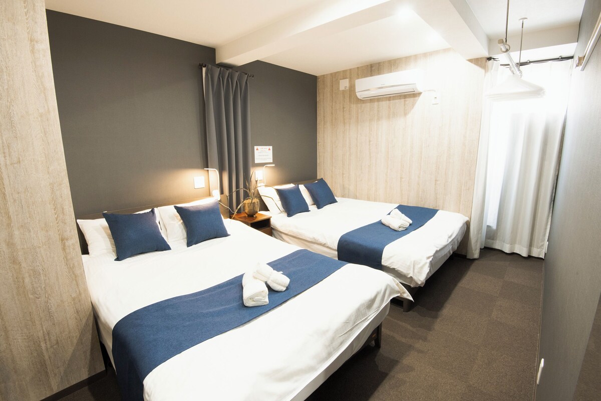 Habitación de hotel con dos camas, cada una con cubrecama azul y ropa blanca. Se observa un mesita de noche con lámpara, una ventana con cortinas y un equipo de aire acondicionado en la pared.