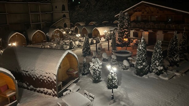 A snow-covered courtyard with several wooden chalets, a central seating area, and a large building with a balcony. - Gasthof Rafting Alm (Haiming)