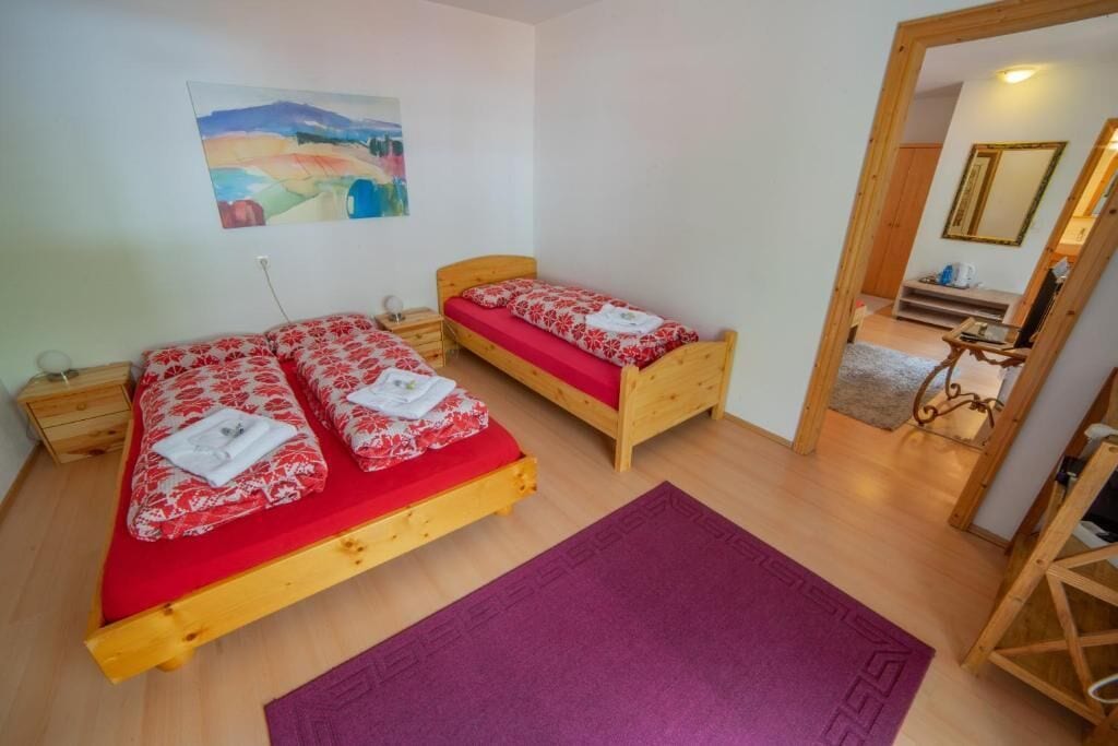 Una habitación con dos camas de madera, ropa de cama roja, una alfombra morada, una mesita de noche de madera y un cuadro en la pared.