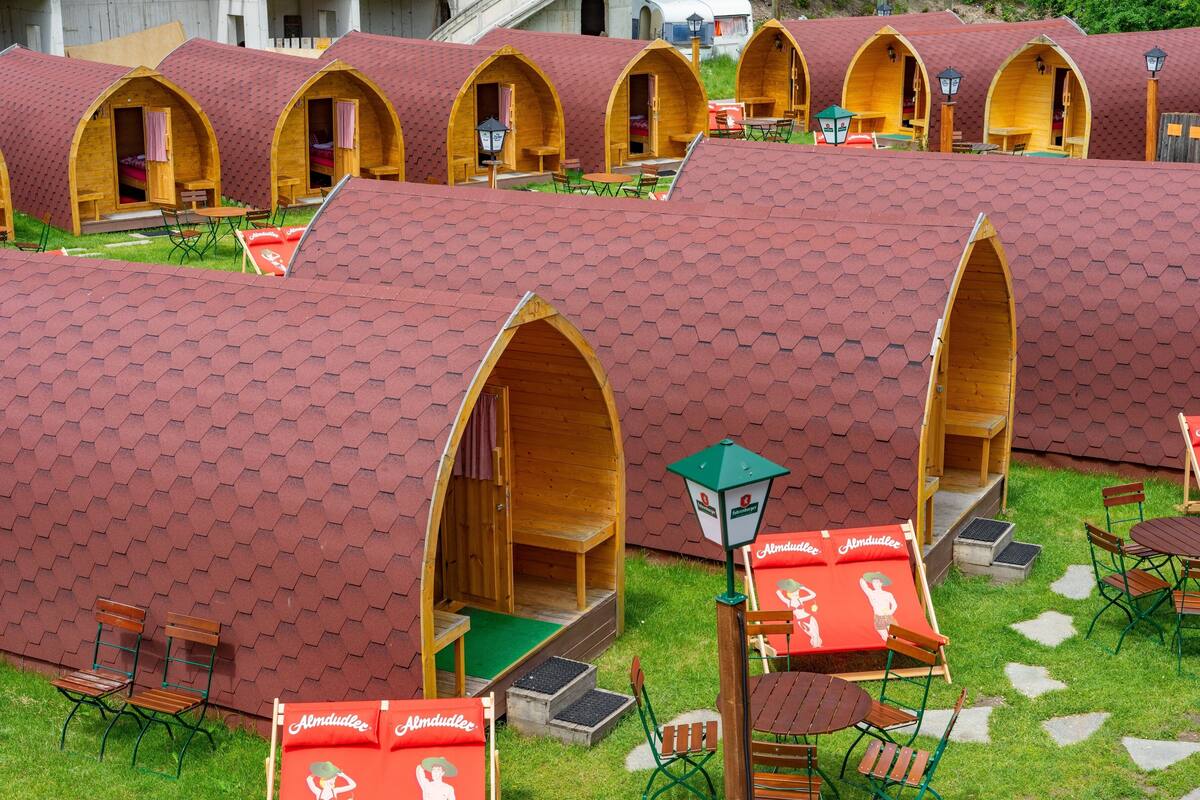 A row of wooden arched tents with green grass in front, each with a small table and chair.