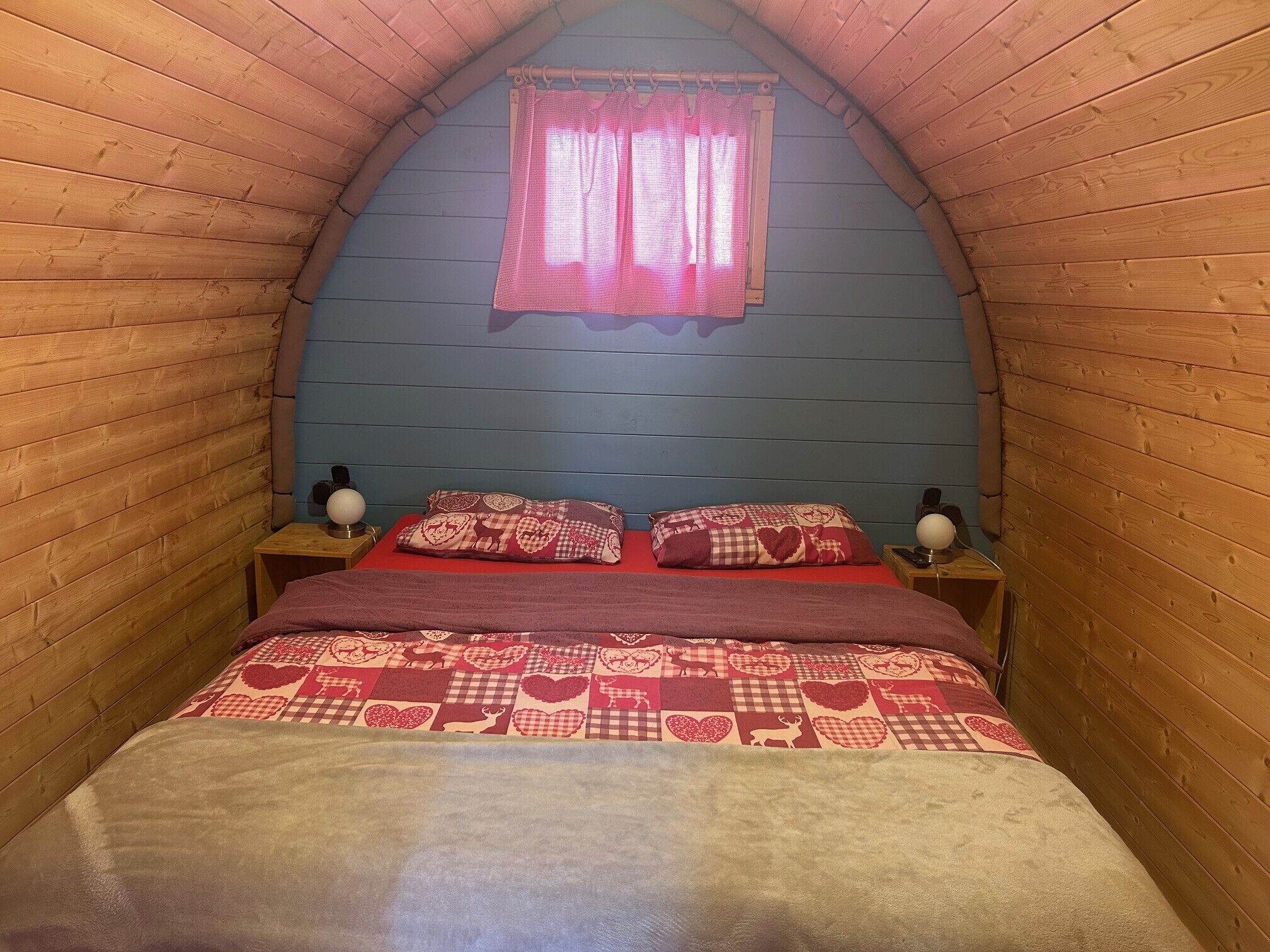 Une pièce étroite et chaleureuse aux murs en bois, avec un plafond en pente, un lit recouvert d’une courtepointe à motifs, deux lampes de chevet et une fenêtre avec des rideaux.