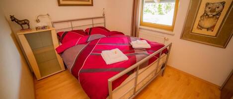 Una cama individual con ropa de cama roja y gris, un aparador de madera y una ventana con cortinas.
