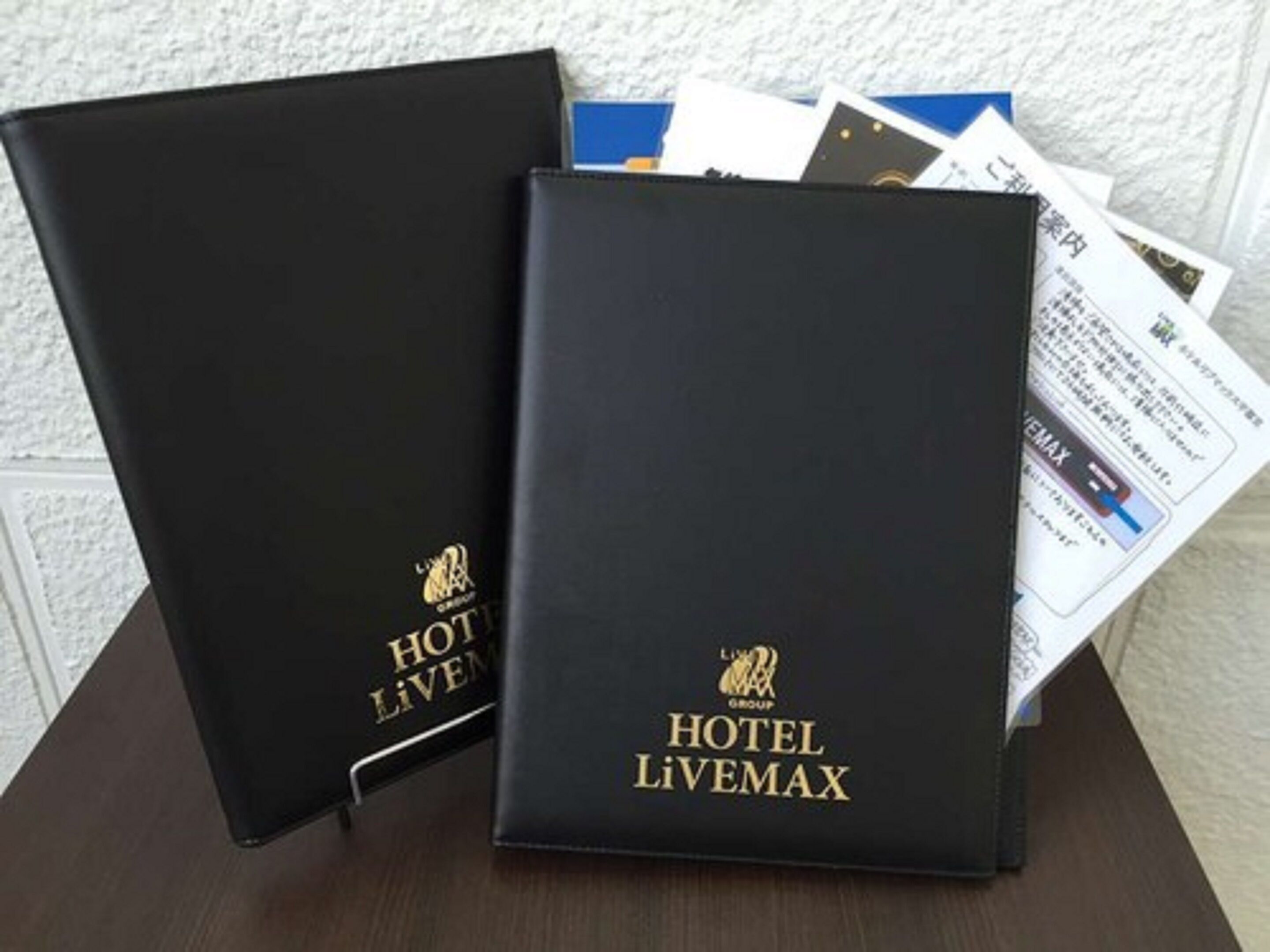 Two black leather-bound books with 'HOTEL LIVEMAX' branding, placed on a wooden surface with some papers partially visible.