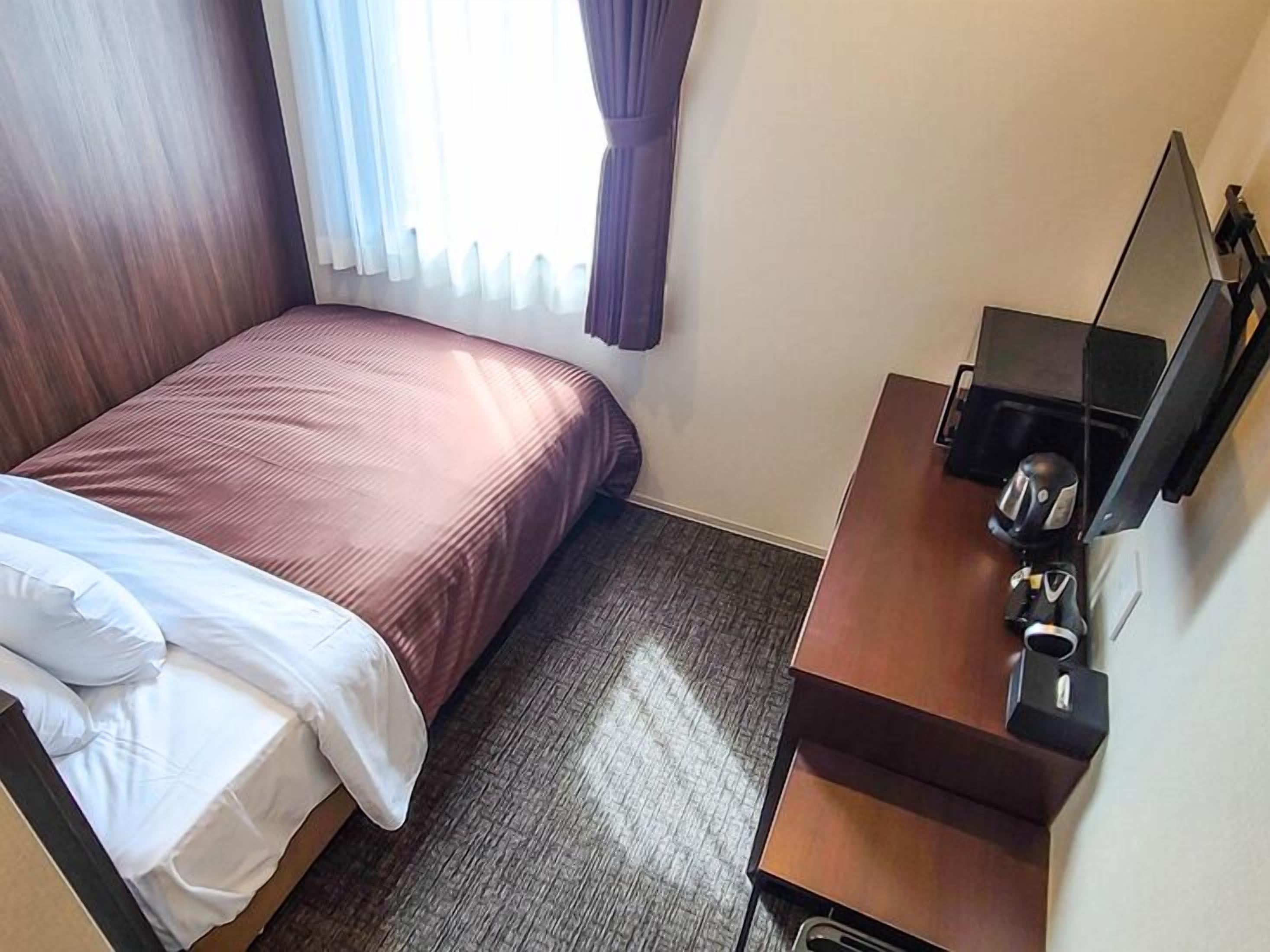 A hotel room with a bed, a desk with a chair, a mirror, and a window with curtains.