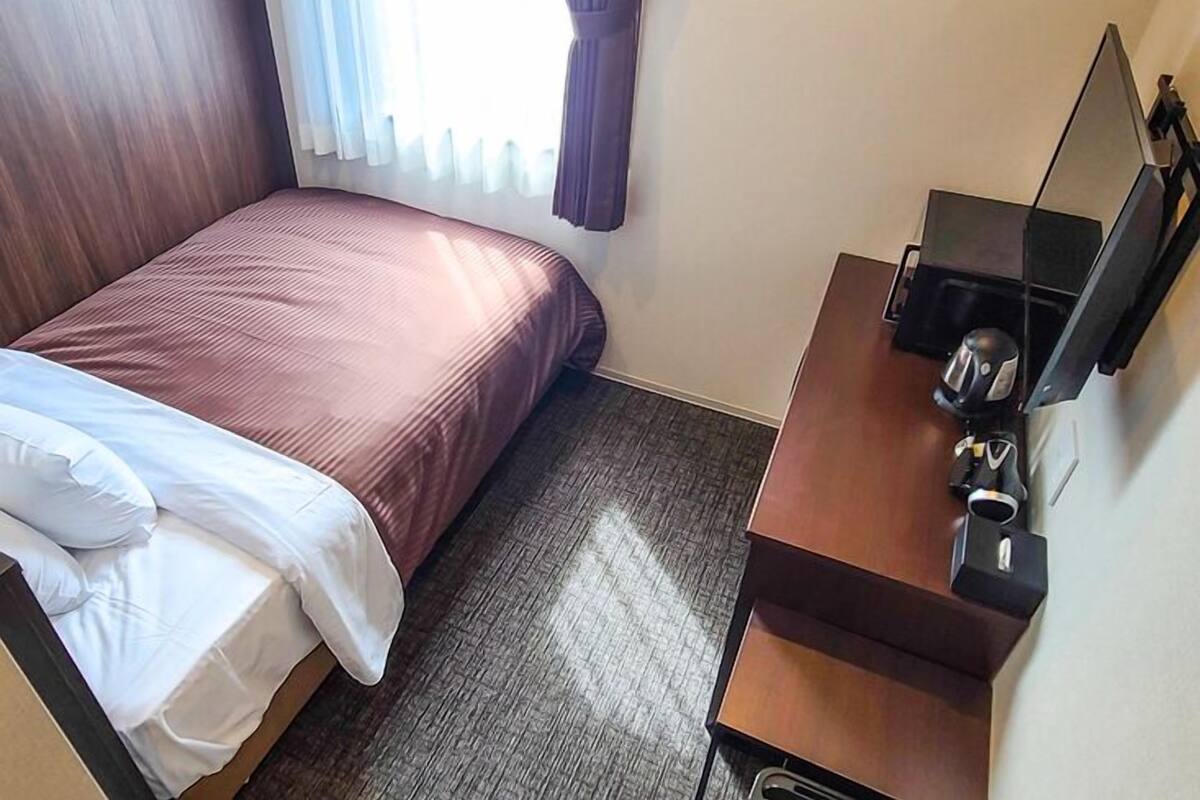 Habitación de hotel con cama, escritorio con silla, espejo y ventana con cortinas.