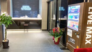 Ein Hotel-Empfangsbereich mit modernem Design, inklusive einer Rezeption, einer Pflanze und einem Automaten mit der Aufschrift "LIVE MAX".