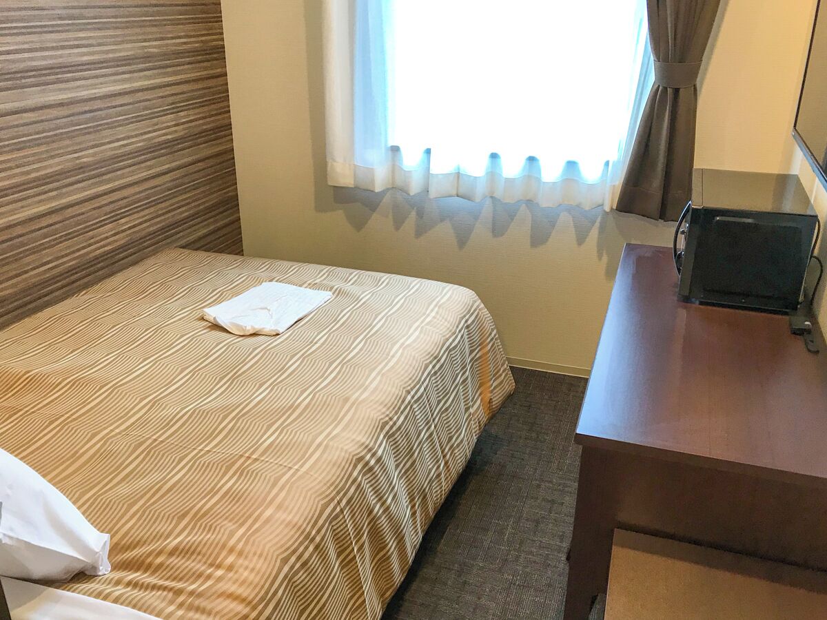 A hotel room with a bed, a bedside table, a window with curtains, and a wall with a textured pattern.