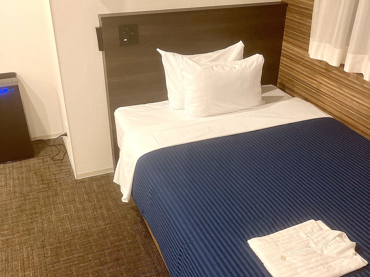 A hotel room with a bed, a headboard, a white pillow, a blue bedspread, and a folded white towel on the bed.