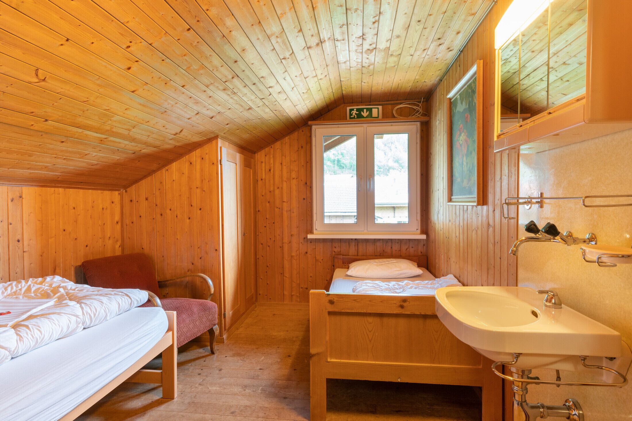Una habitación de cabaña de madera con una cama, una silla, un ventanal pequeño y un lavabo.