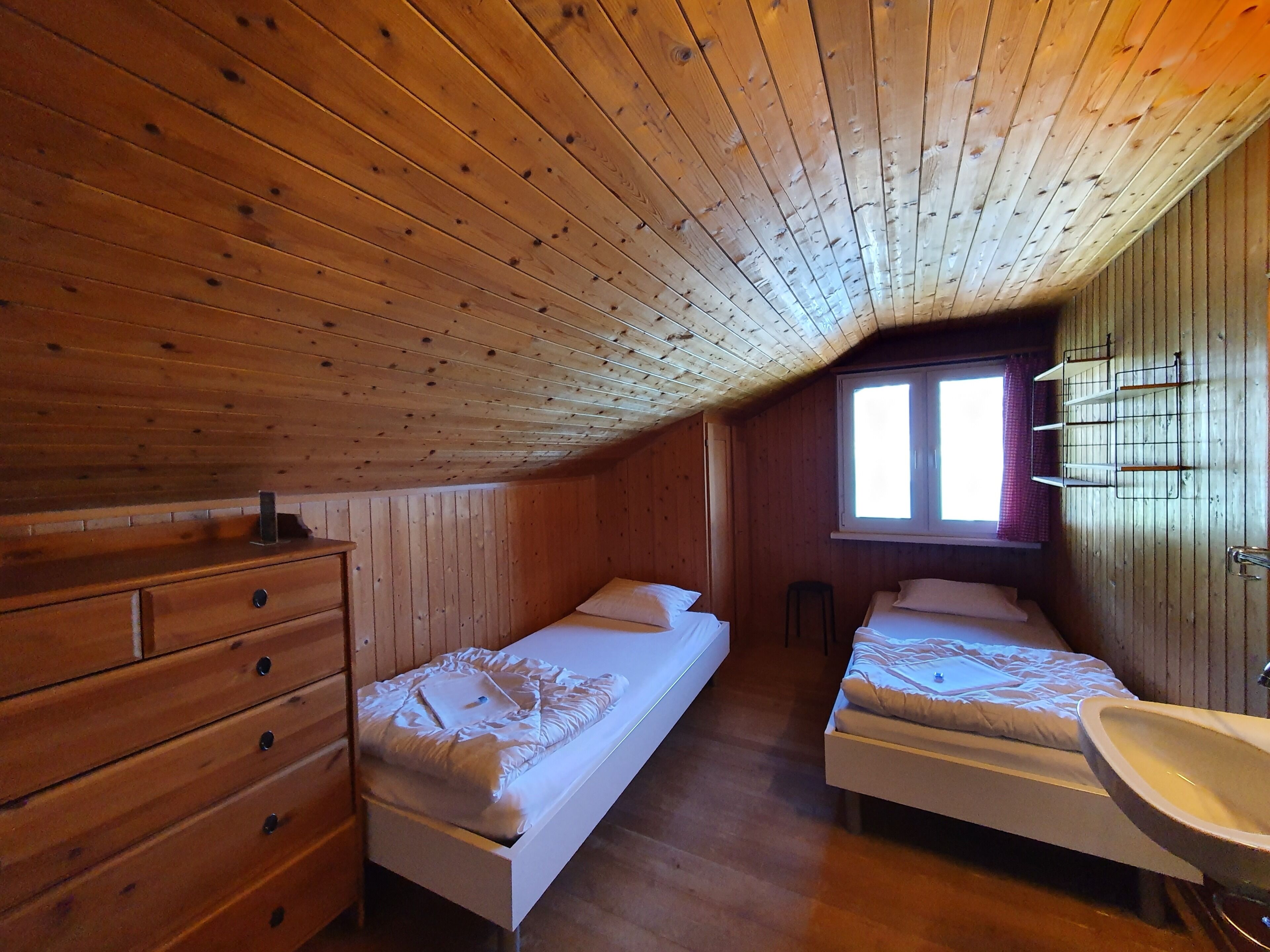 A small, wooden cabin with two single beds, a wooden dresser, a window with curtains, and a sink.