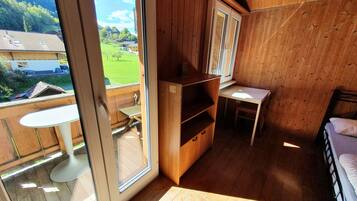 Una habitación de madera con una cama, un escritorio y una silla. Se ve un paisaje verde y un edificio a través de una ventana.