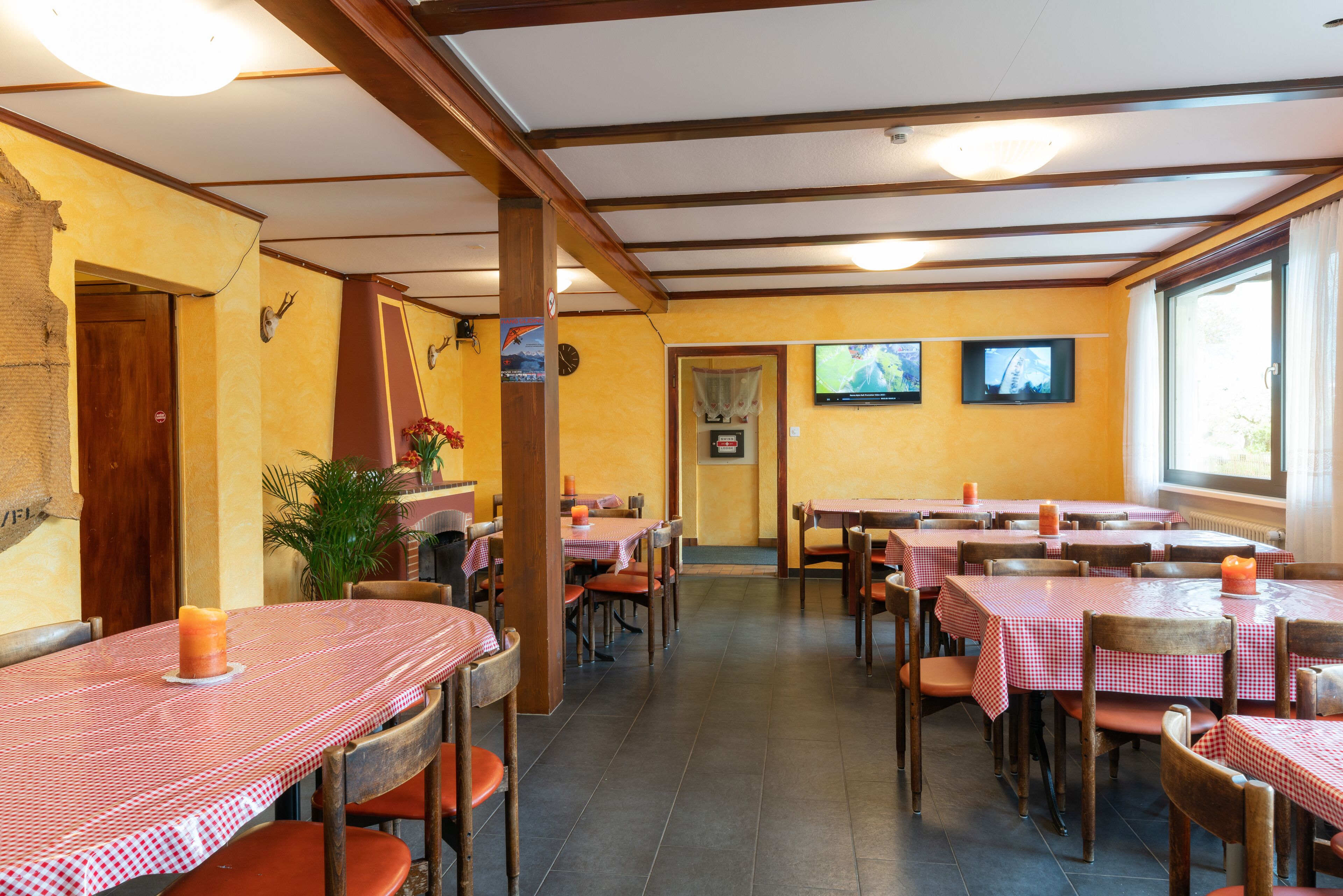 A restaurant with multiple tables and chairs, wooden beams on the ceiling, and a large window on the right.