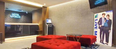 Un vestíbulo de hotel con diseño moderno, que presenta una zona de estar roja, un quiosco digital de información y una gran pantalla con el nombre del hotel.