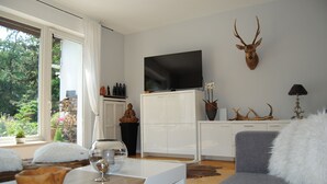 A modern living room with a large flat-screen TV, a mounted deer head, and a view of greenery outside. - Exclusive villa with garden, sauna and forest access  (Solingen)