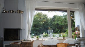 A cozy living room with a fireplace, wicker furniture, and a view of a garden through a large window. - Exclusive villa with garden, sauna and forest access  (Solingen)