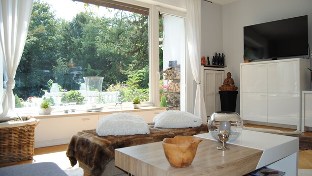 A modern living room with a large window, a white sofa, a wooden coffee table, and a television on a cabinet. - Exclusive villa with garden, sauna and forest access  (Solingen)