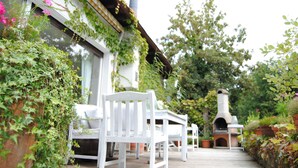 A patio with white chairs and a brick oven, surrounded by greenery and potted plants. - Exclusive villa with garden, sauna and forest access  (Solingen)