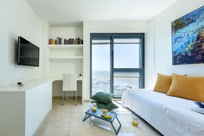 Una habitación de hotel moderna con una cama, un escritorio, una silla, una mesita con productos para el desayuno, una estantería y un ventanal con vistas al puerto deportivo.