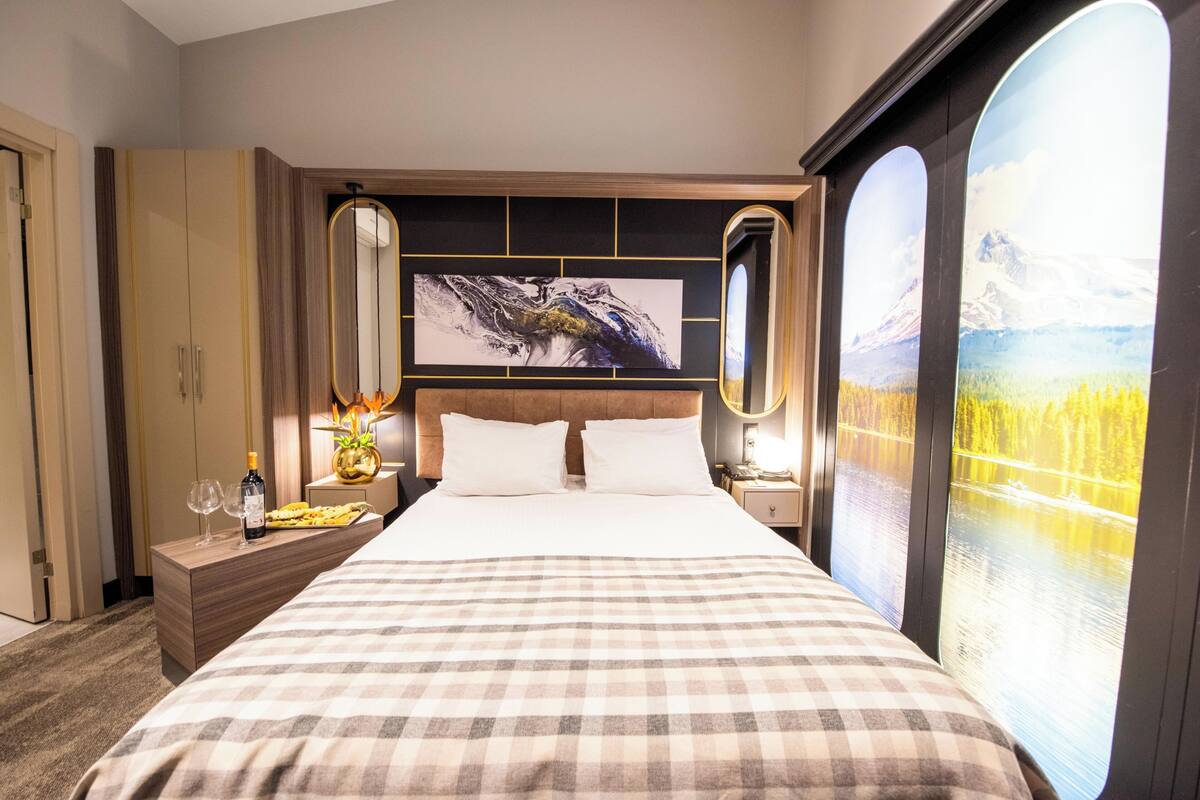 A hotel room with a large bed, a nightstand with a bottle and glasses, a wall-mounted painting, and a window with a view of a lake and mountains.