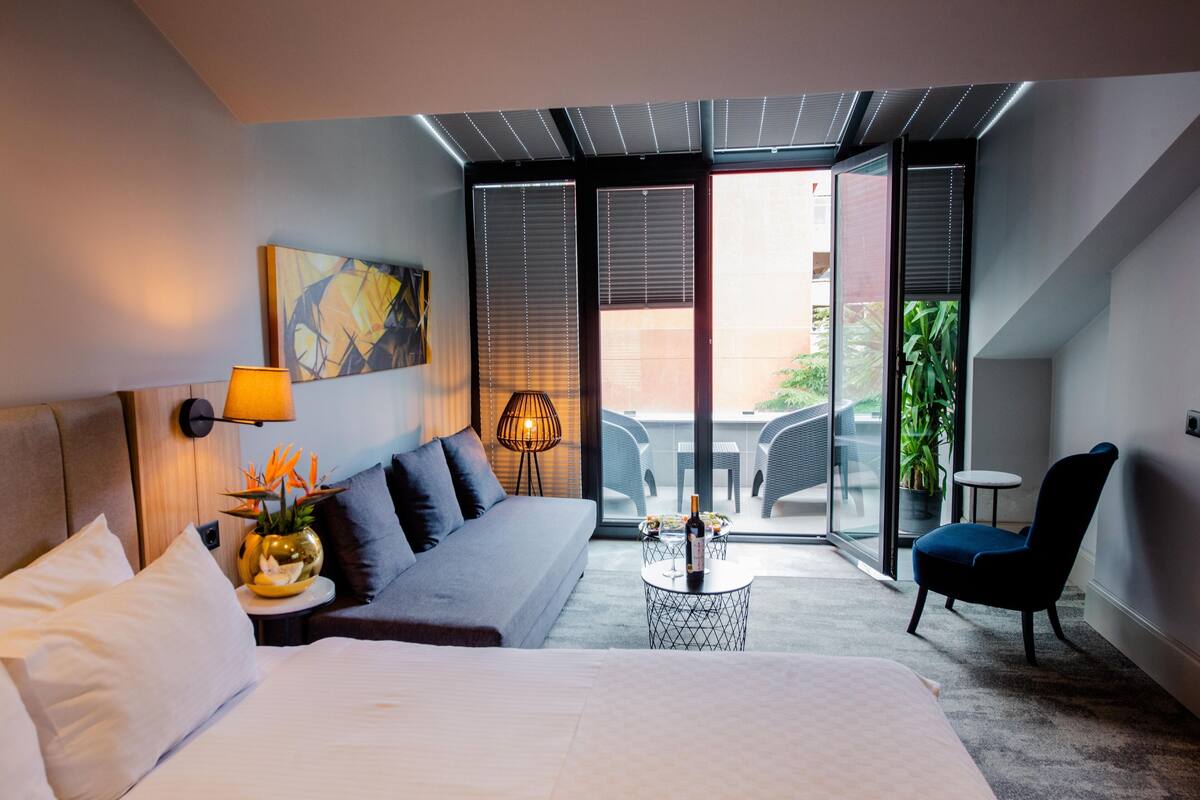 A modern hotel room with a bed, sofa, lounge chair, and a balcony with a view.