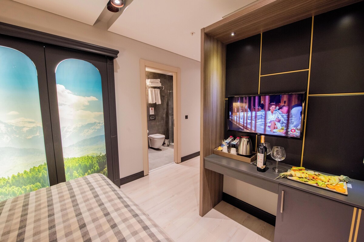 A modern hotel room with a large bed, a television, a small kitchen area with a breakfast tray, and a scenic mural on the wall.