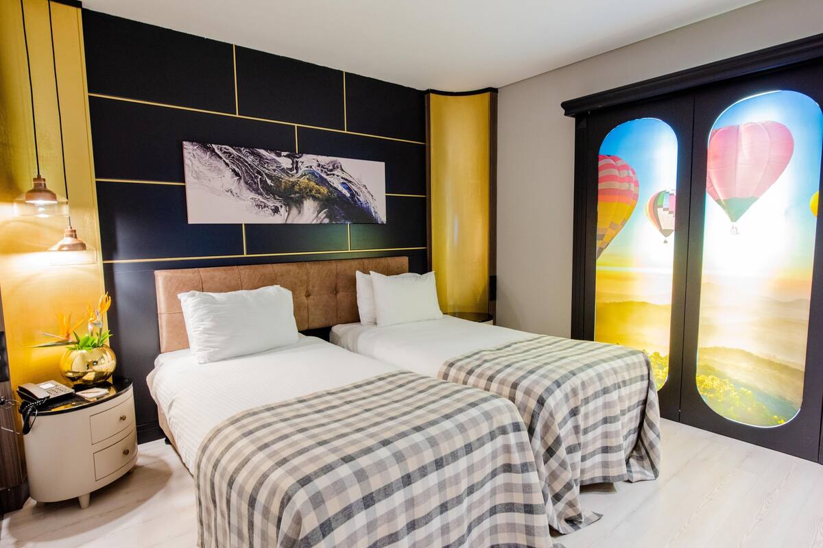 A hotel room with two beds, a nightstand, a lamp, a painting, and a view of hot air balloons.