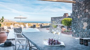 Un patio exterior con una pared de piedra, una mesa con una fuente de uvas y vasos, y vistas al océano.