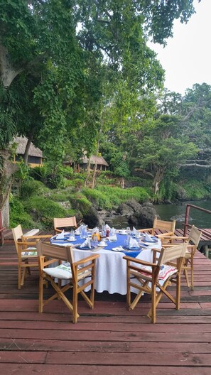 An outdoor dining area with a table set for four, surrounded by wooden chairs, overlooking a body of water and lush greenery. - Mfangano Island Lodge (Mfangano Island)
