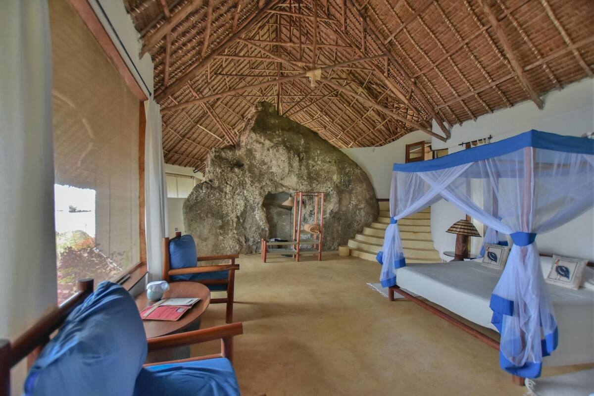 A room with a thatched roof, a bed with a mosquito net, a wooden chair, and a table with a notebook and pen.