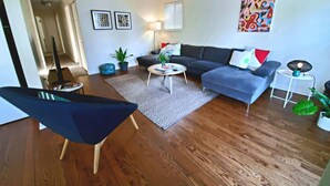 Ein modernes Wohnzimmer mit einem dunkelblauen Sessel, einem grauen Ecksofa, einem runden Couchtisch und einem gemusterten Teppich.