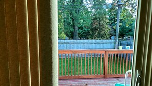 Blick von einem Zimmer auf eine Holzterrasse mit Metallgeländer, einem grünen Stuhl und einem Holzzaun im Hintergrund.