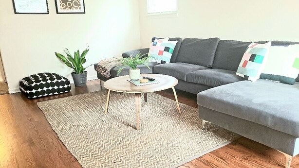 Ein Wohnzimmer mit grauem Sofa, einem runden Couchtisch, einem gemusterten Teppich und zwei Topfpflanzen.