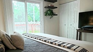 Ein Schlafzimmer mit Bett, Kissen, einem Fenster mit Vorhängen, einer Pflanze auf einem Regal und einem Fernseher auf einem Schrank.
