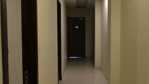 A long, narrow hallway with a dark wooden door on the left, a light-colored tiled floor, and a dark door at the end. - Hotel KK Continental (Amritsar)