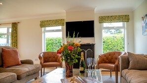 A living room with a sofa, armchairs, a coffee table with a magazine, a vase with flowers, a bottle of champagne, and a fireplace. - Wonderful Spacious Family Home in Constantine Bay Situated on Road to Beach (Constantine Bay)