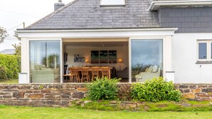A house with a stone wall, a dining area with a table and chairs, and a large sliding glass door leading to an interior space. - Wonderful Spacious Family Home in Constantine Bay Situated on Road to Beach (Constantine Bay)