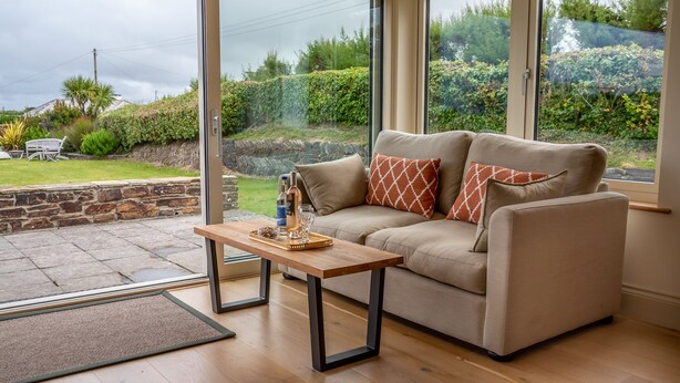 A cozy living room with a beige sofa, two patterned cushions, a wooden coffee table, and a glass door leading to a garden. - Wonderful Spacious Family Home in Constantine Bay Situated on Road to Beach (Constantine Bay)