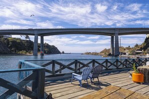 Une terrasse en bois avec des chaises et un pot de fleurs, donnant sur une étendue d’eau et un pont.