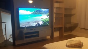 Un dormitori amb una televisió que mostra una escena natural, un llit amb una tovallola enrotllada i un moble prestatge de fusta.