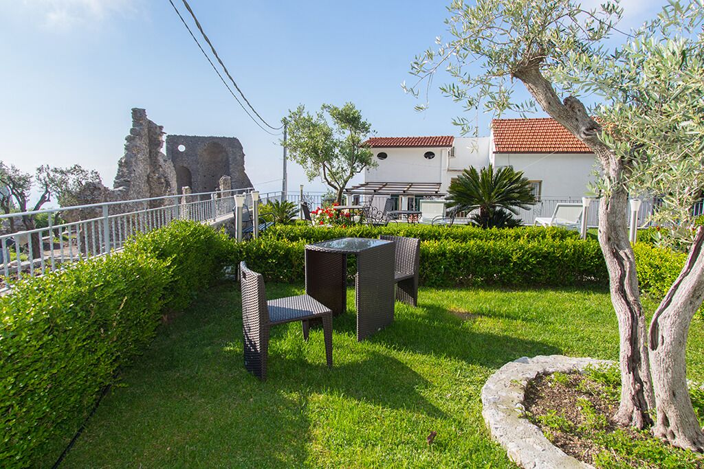 A garden with a table and chairs, a white building with a red roof, and a stone structure in the background.