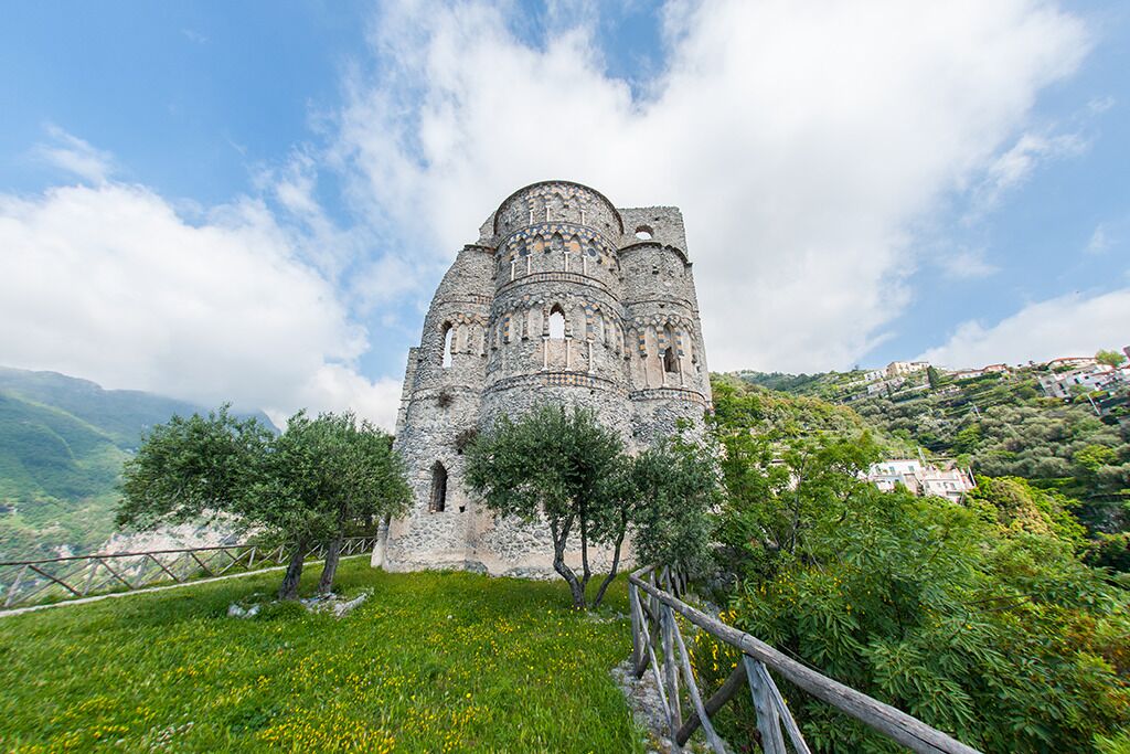 Un bâtiment historique en pierre, surmonté d’un dôme, entouré d’une végétation luxuriante et adossé à un paysage montagneux.