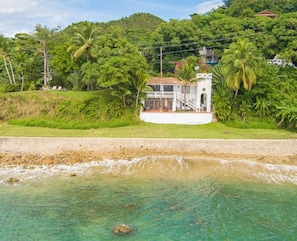 A coastal property with a white building, a staircase leading to a balcony, and lush greenery.