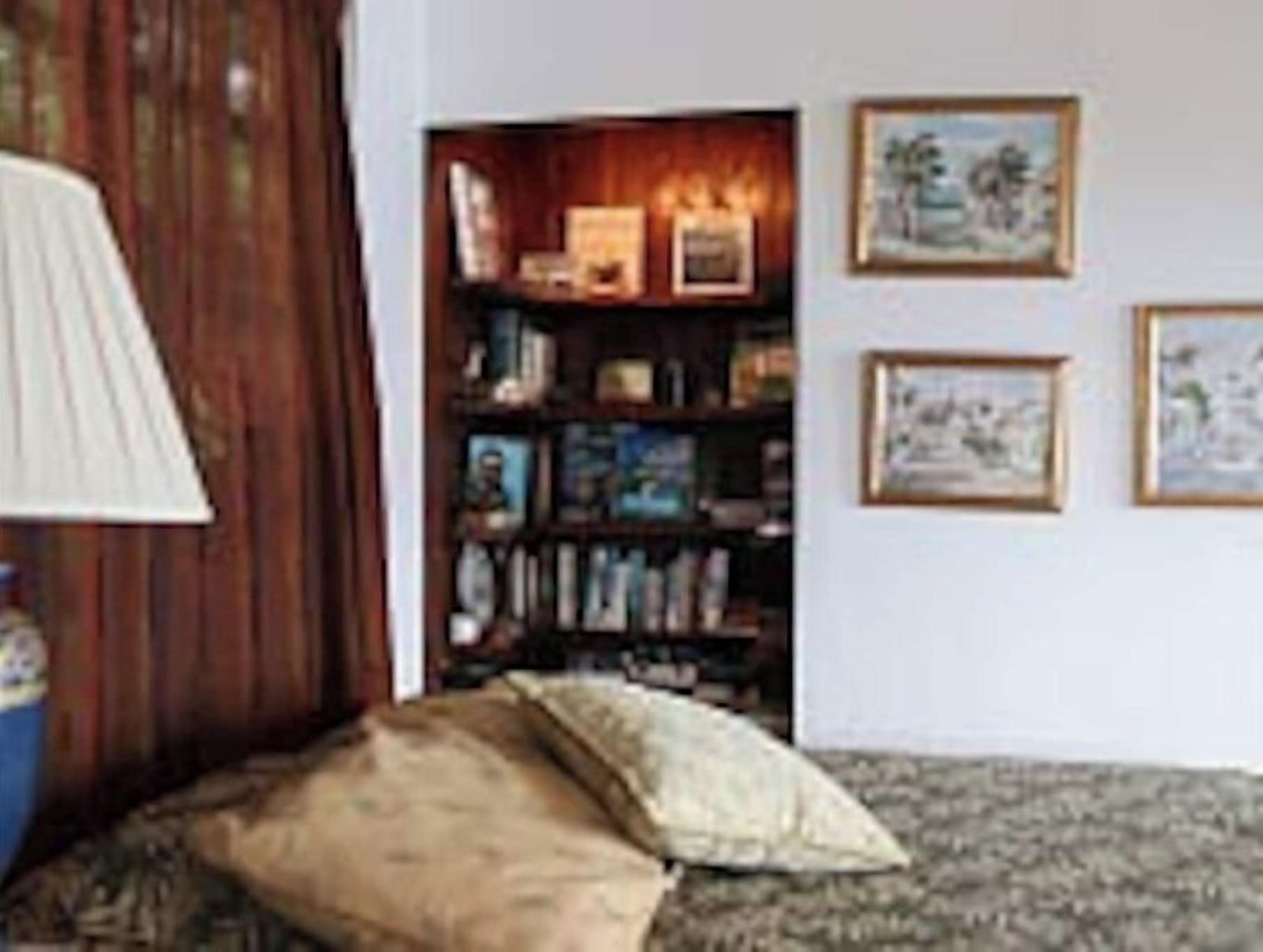 Een kamer met een houten vloer, een bank, een lamp en een boekenkast met ingelijste foto's.