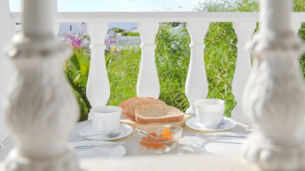 Une table dressée avec de la vaisselle blanche, du pain et un petit bol, avec vue sur la verdure et une balustrade blanche.