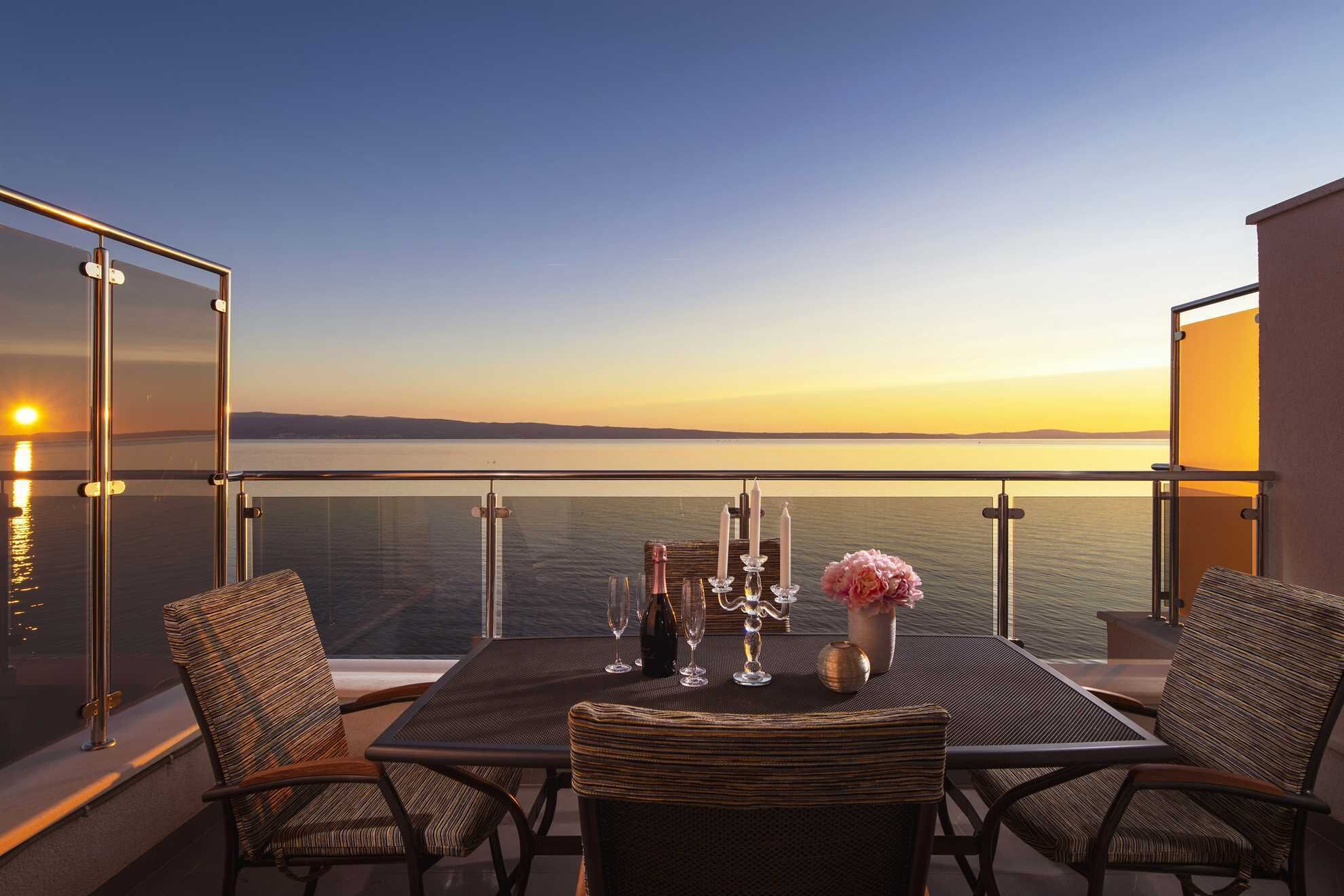 A balcony with a table set for two, overlooking a calm body of water during sunset.