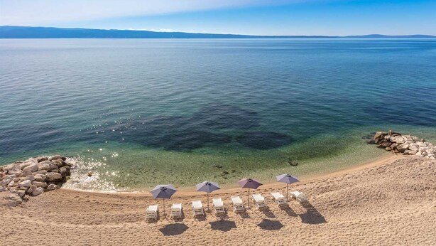 A beach with clear water, a rocky shoreline, and several sun loungers with umbrellas. - Tanita Beachfront Penthouse (Podstrana)